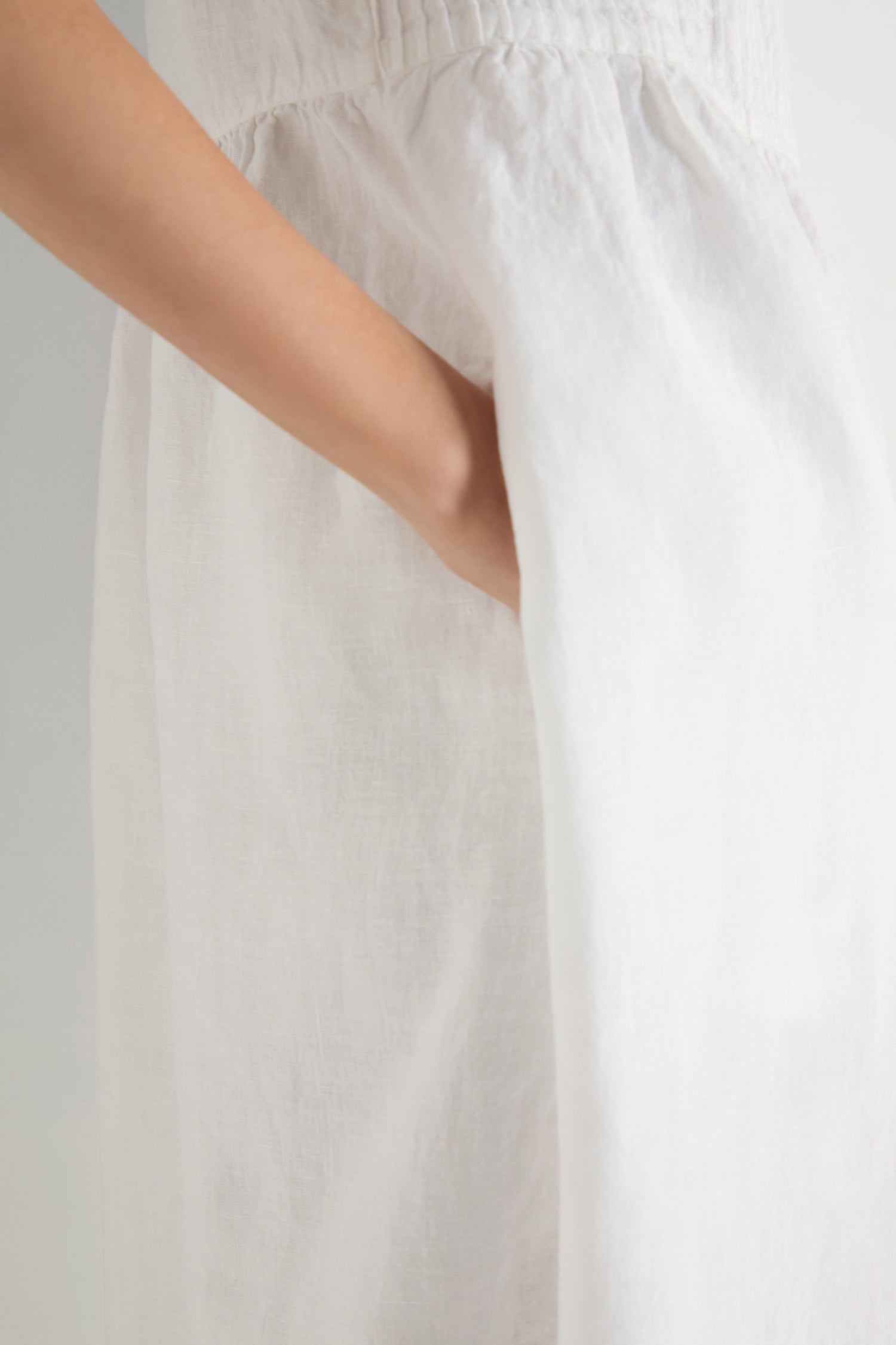 A person wearing a white Pleat Front Linen Bubble Dress has their hand in the dress pocket, shown from the shoulder down against a plain background.
