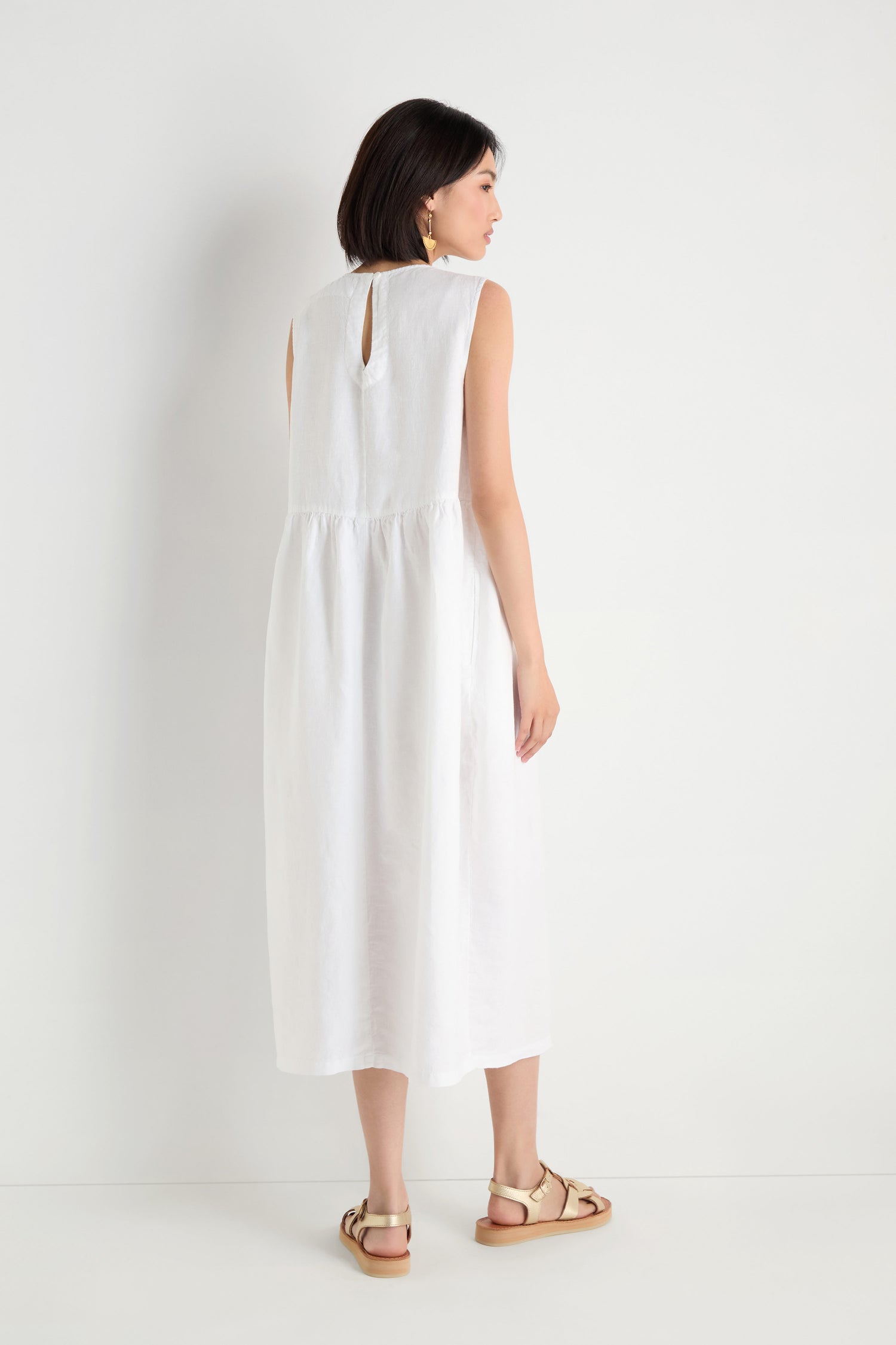 Woman standing against a white wall, facing away, wearing a sleeveless Pleat Front Linen Bubble Dress and beige sandals.