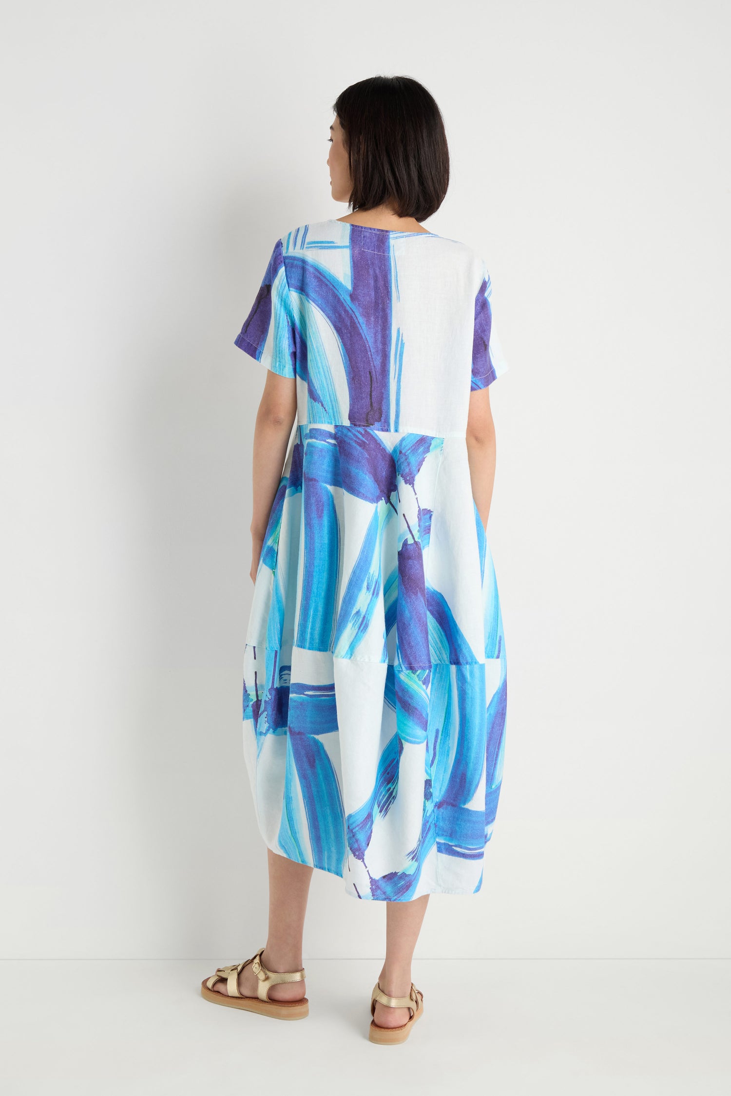 Back view of a woman in the Brushstroke Linen Bubble Dress, featuring a blue and white print, short sleeves, and a full skirt, paired with gold flat sandals.