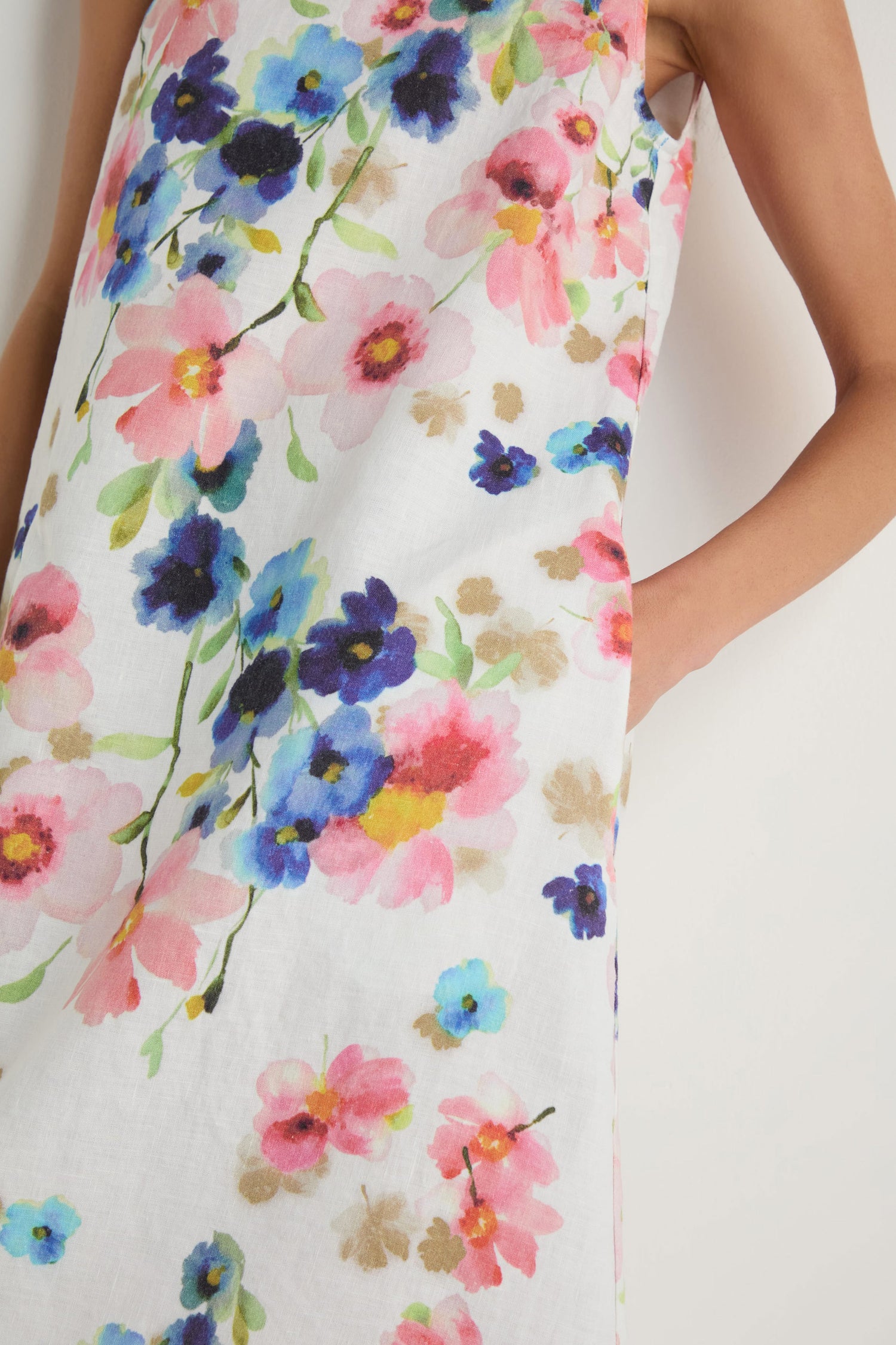 Wearing the Primrose Print Sleeveless Linen Dress, a person stands with one arm bent and a hand in the pocket, showing off its colorful print.