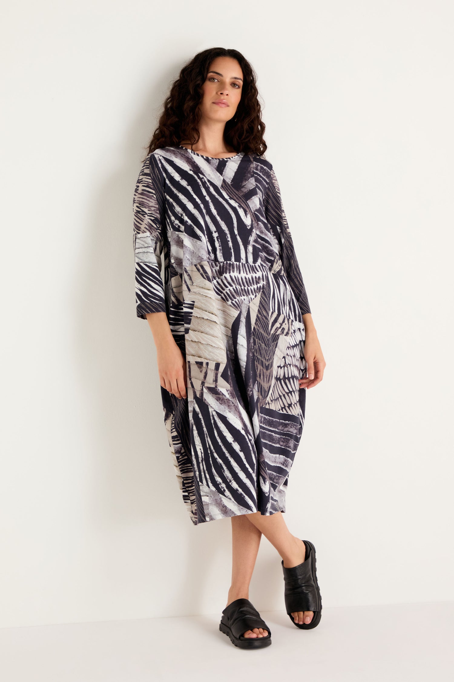 A woman stands against a plain white background wearing the Monochrome Patchwork Jersey Dress—a loose, knee-length dress featuring a black and white abstract pattern, three-quarter sleeves, and paired with black open-toe sandals.
