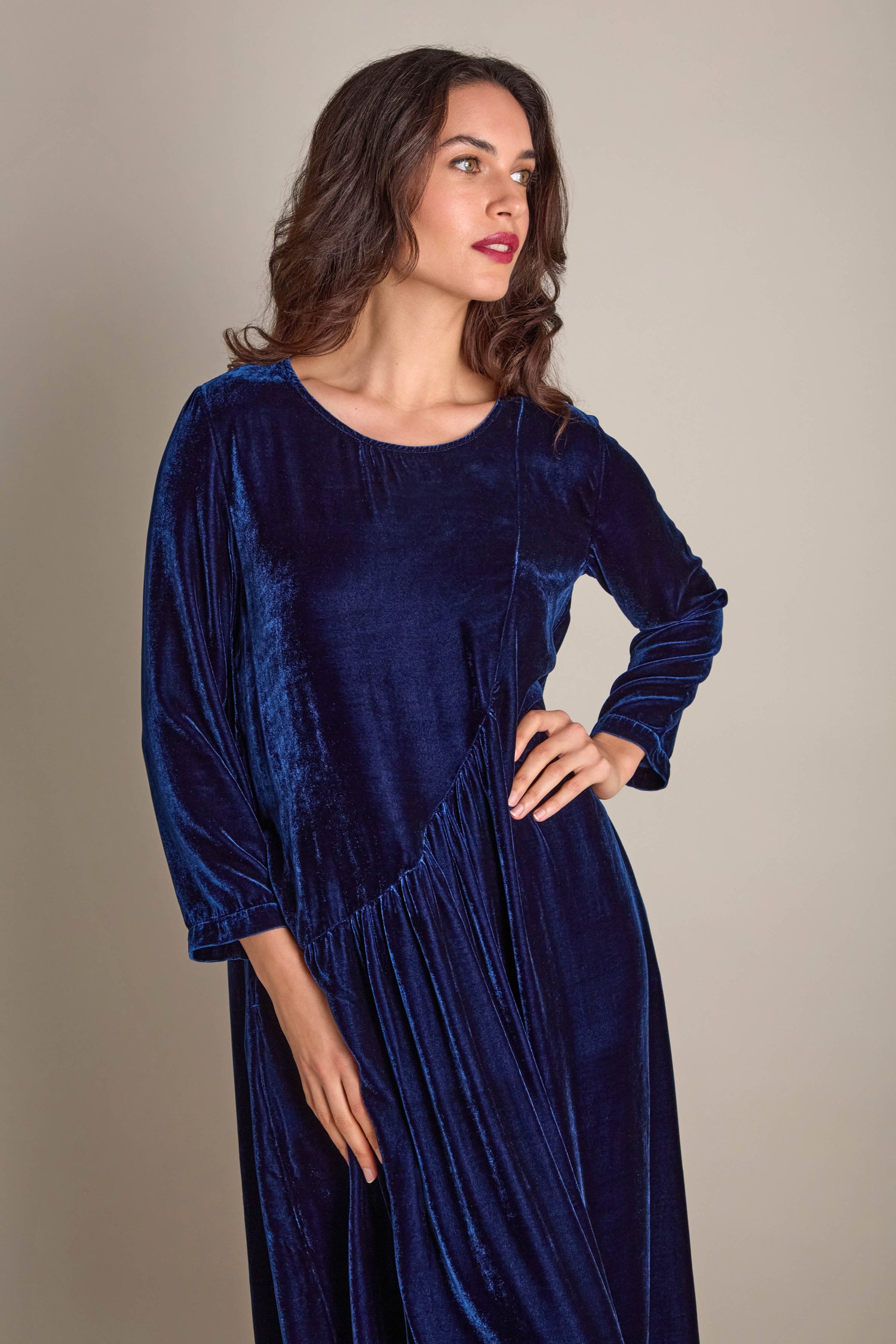 A woman poses elegantly in the Drape Front Silk Velvet Dress, showcasing its long sleeves and asymmetric draped front in a deep blue hue, radiating luxurious elegance against a simple backdrop.