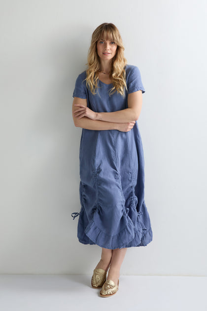 A woman stands against a white wall, wearing the Ruched Hem Dress in loose blue crinkled linen with gold woven flats and a neutral expression—ideal for versatile styling.