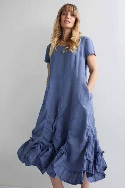 A woman stands against a plain light-colored wall wearing the Ruched Hem Dress, a loose-fitting, short-sleeved blue linen dress featuring subtle ruche details near the hem.