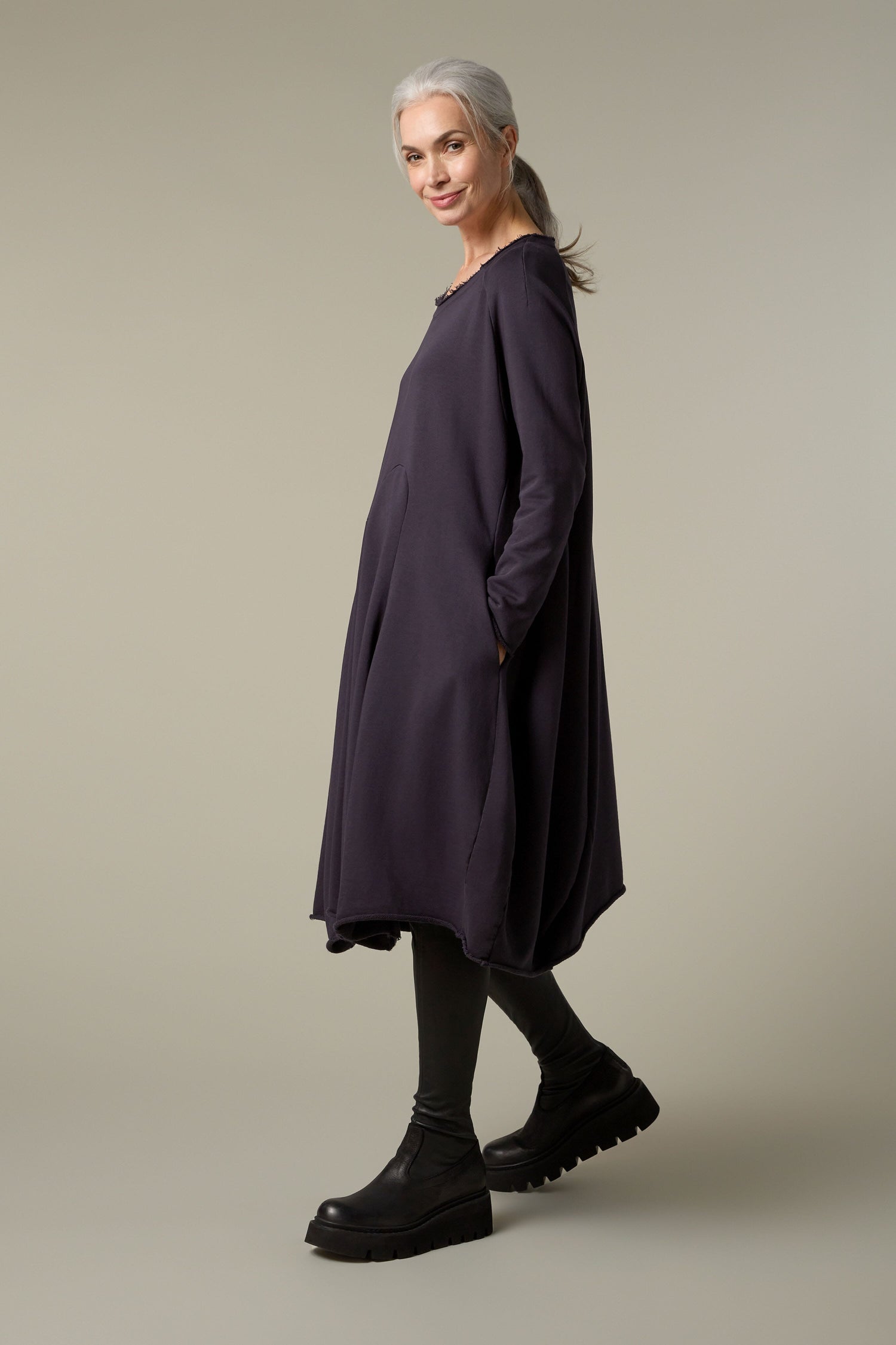 A woman wearing a Godet Jersey Dress in a purple color, paired with black leggings made of weighty jersey.