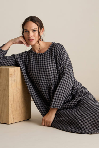 A person wearing the Linen Check Big Pocket Dress, made from soft linen fabric with a black and white checkered pattern, sits on the floor leaning against a wooden block with their head resting on their hand. The relaxed tunic style of the dress exudes a comfortable vibe.