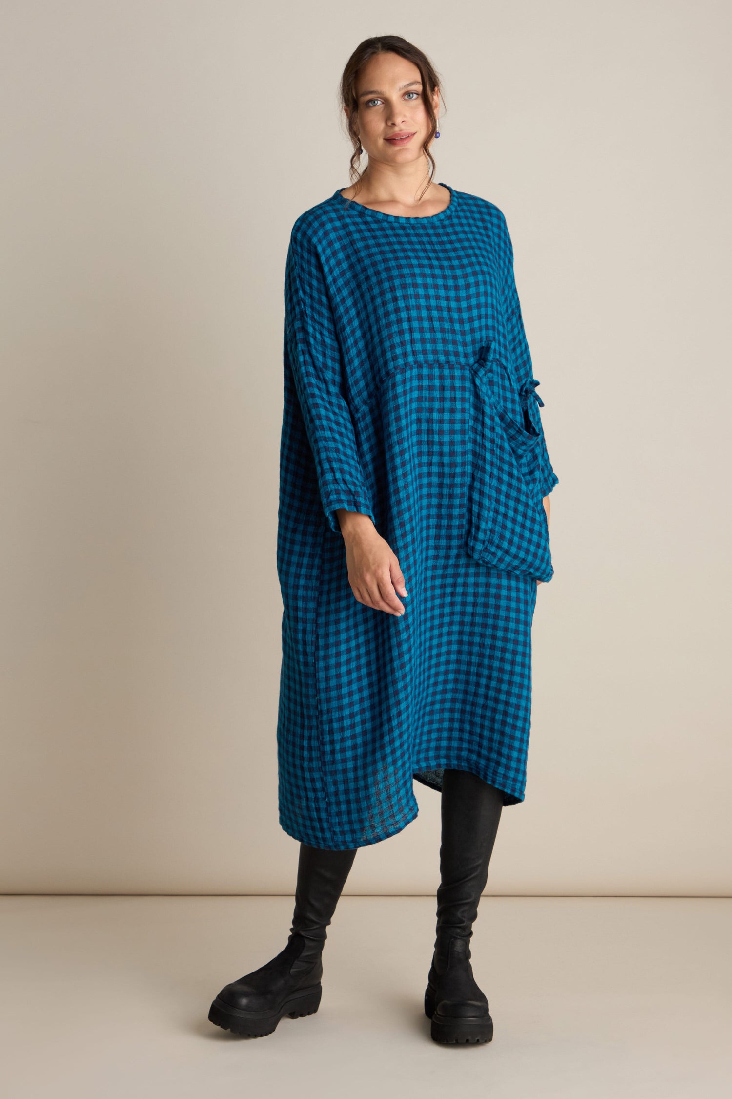 Person standing against a plain background, wearing the Linen Check Big Pocket Dress with its relaxed tunic style and oversized feature pocket, paired with black leggings and chunky black boots.