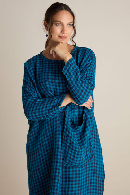 A woman in a relaxed tunic style Linen Check Big Pocket Dress, in blue and black checkered pattern, with one hand resting on her chin and the other holding a matching checkered tote bag.