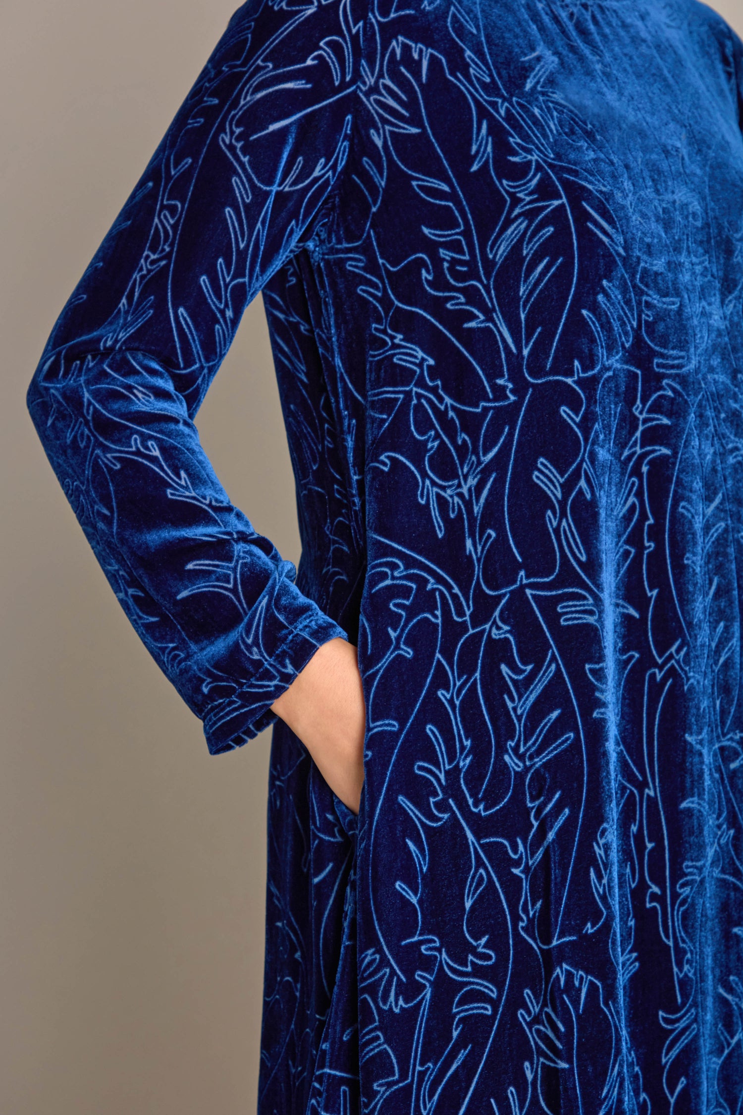 A person is elegantly dressed in the Vine Devore Dress, a blue velvet gown with a vine devoré design, casually resting their right hand in the dress pocket.