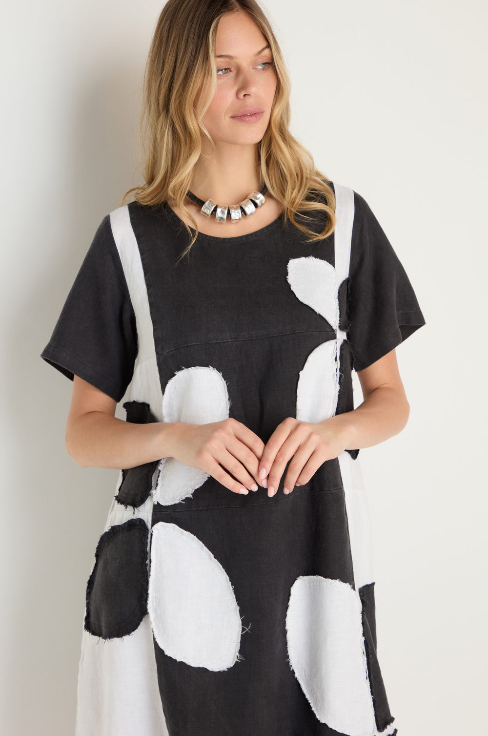 A woman with long blonde hair models the Quant Monochrome Linen Dress, featuring bold black and white abstract shapes, paired with a chunky silver necklace against a simple light backdrop.