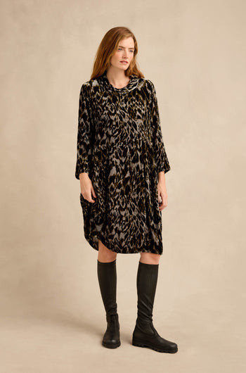 A woman stands against a plain background, wearing the Animal Print Cowl Neck Velvet Dress—a loose, knee-length velvet dress featuring an eye-catching animal print, long sleeves, and a stylish cowl neck—paired with black knee-high boots.