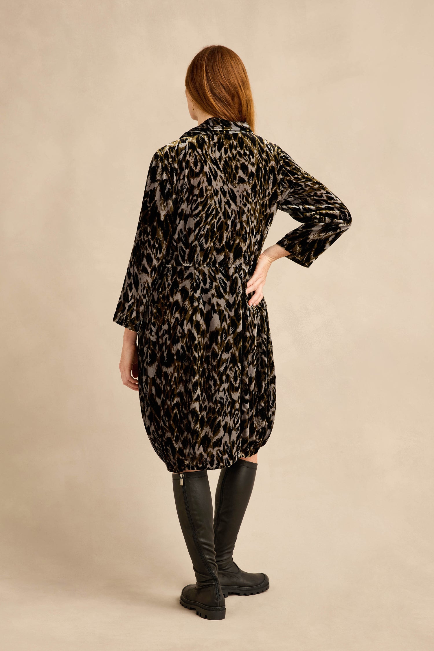 A person with long auburn hair stands facing away, wearing the Animal Print Cowl Neck Velvet Dress—loose-fitting and knee-length—paired with black knee-high boots against a plain beige background.