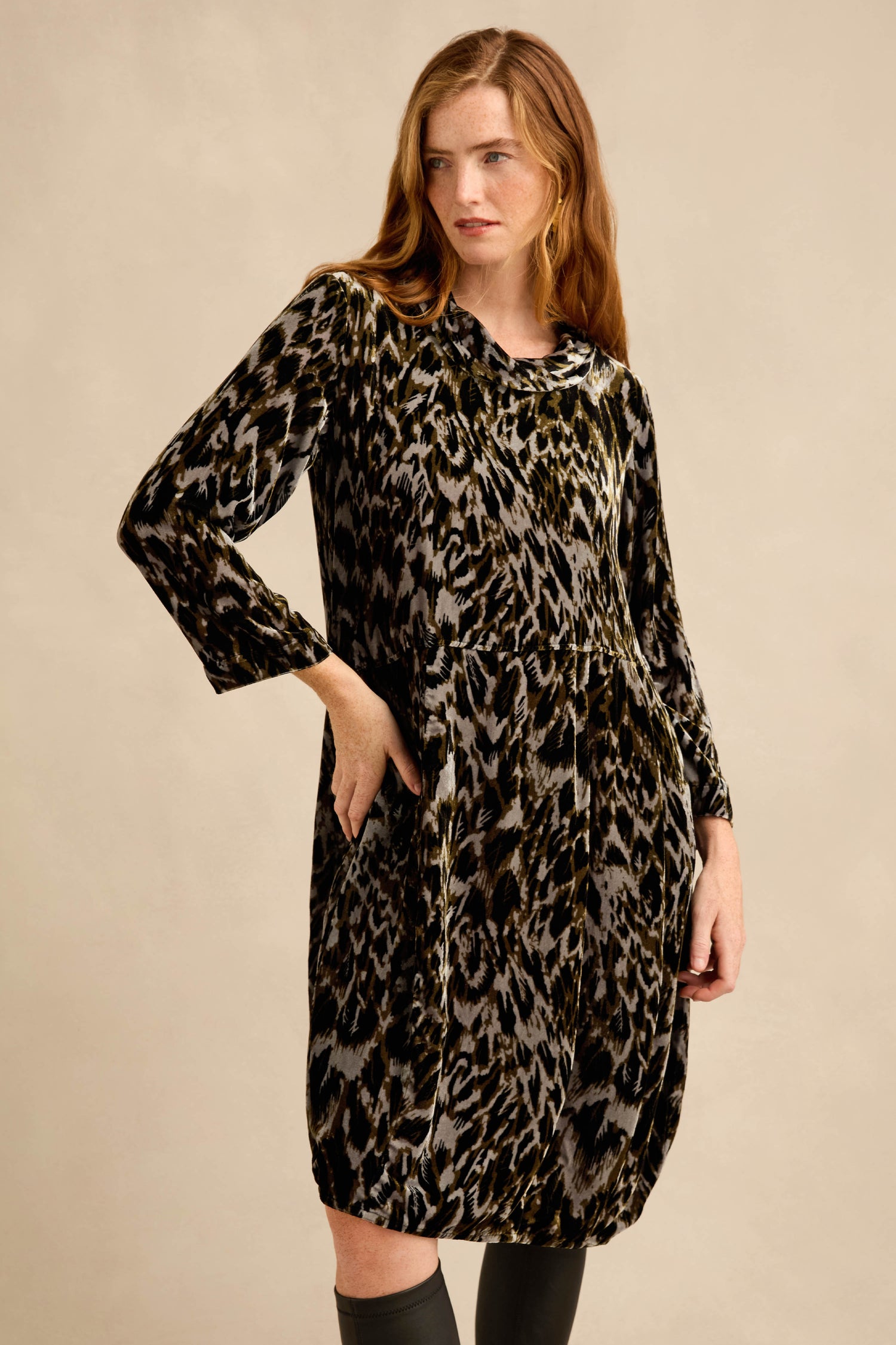 A woman stands against a beige background with one hand in her pocket, wearing the Animal Print Cowl Neck Velvet Dress—a long-sleeved, knee-length dress featuring a chic cowl neckline and stylish animal print.