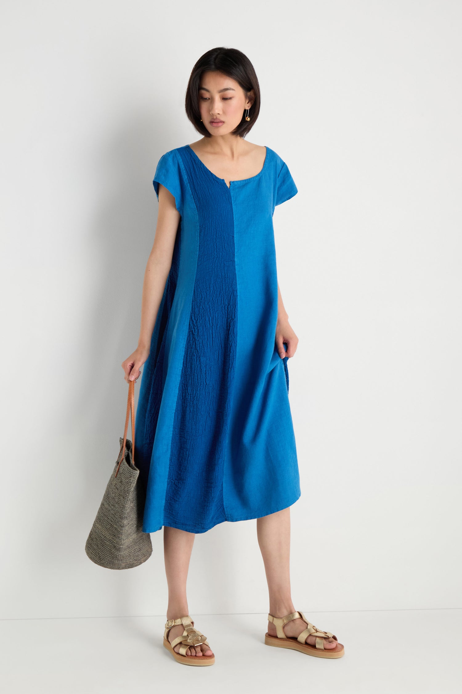 A woman stands against a plain white background wearing the Panelled Linen Flared Dress in knee-length blue with short sleeves, styled with beige sandals and holding a gray tote bag.