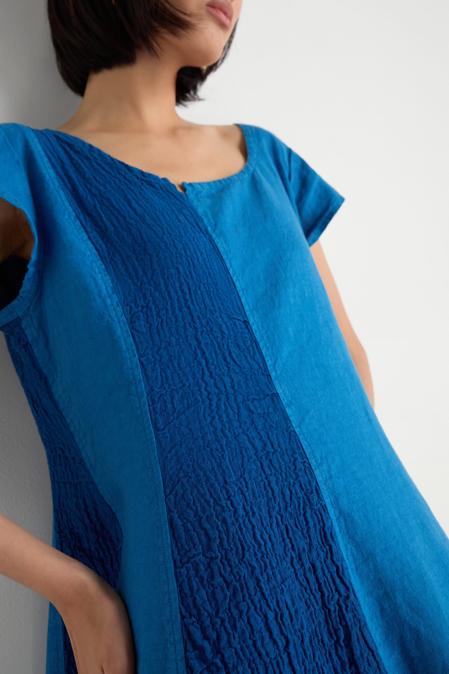 A woman stands against a white wall, hands in her pockets, wearing the Panelled Linen Flared Dress in blue with short sleeves.