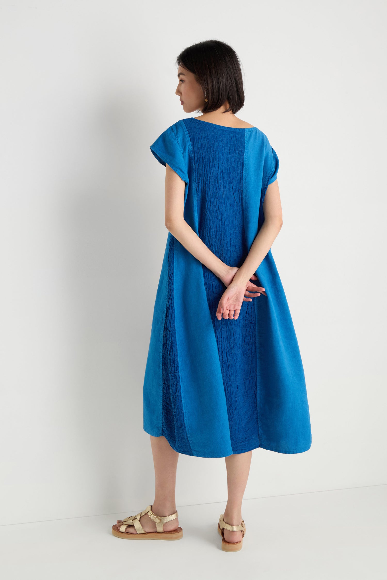 A woman stands facing away against a white background, wearing the Panelled Linen Flared Dress in blue, knee-length, paired with beige sandals and her hands clasped behind her back.