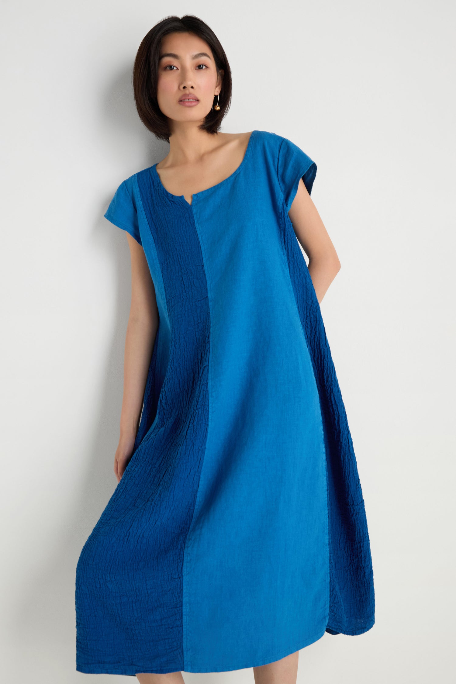 Woman models the Panelled Linen Flared Dress, a knee-length, short-sleeved linen dress featuring a relaxed fit and color-block blue design, set against a plain white background.