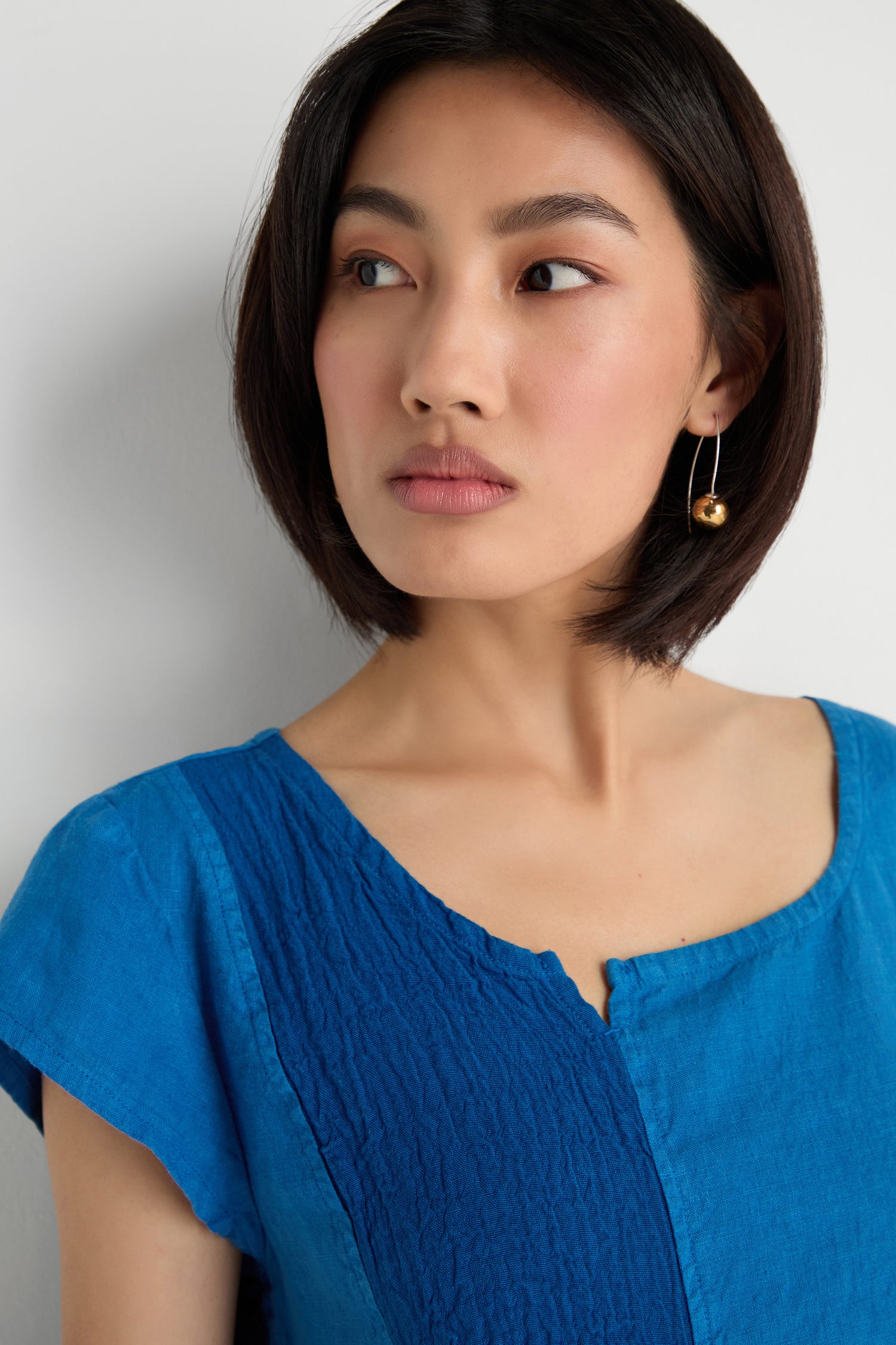 Woman with short dark hair wears the Panelled Linen Flared Dress in blue and gold dangle earrings, looking to the side against a plain light background.
