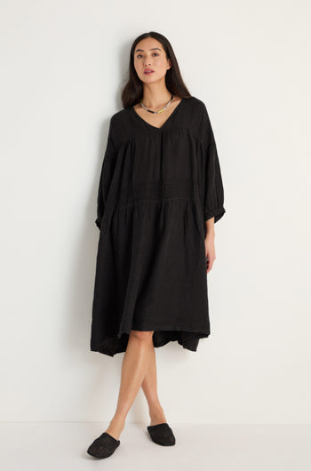A woman stands against a white background wearing the Easy Fit V-Neck Black Linen Dress, styled with black slip-on shoes and a necklace.