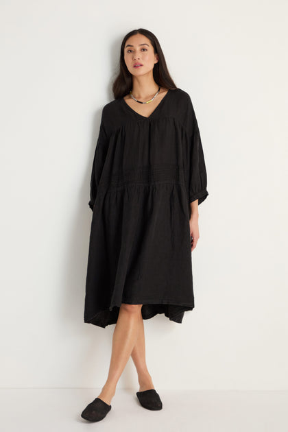 A woman stands against a white background wearing the Easy Fit V-Neck Black Linen Dress, styled with black slip-on shoes and a necklace.