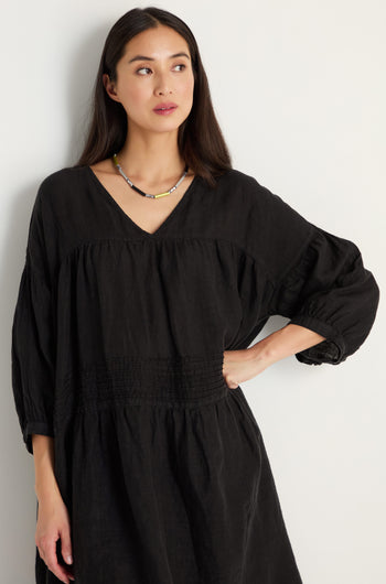 A woman with long dark hair wears the Easy Fit V-Neck Black Linen Dress and a beaded necklace as she stands against a plain light-colored wall.