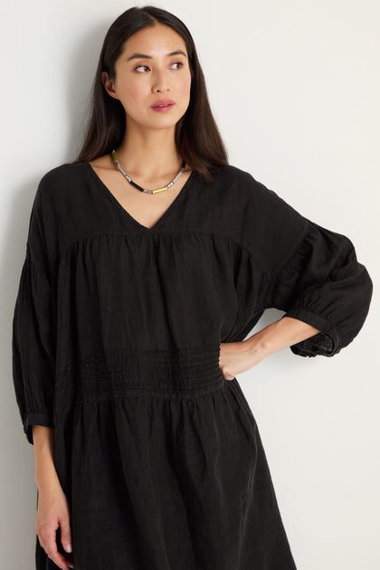 A woman with long dark hair wears the Easy Fit V-Neck Black Linen Dress and a beaded necklace as she stands against a plain light-colored wall.