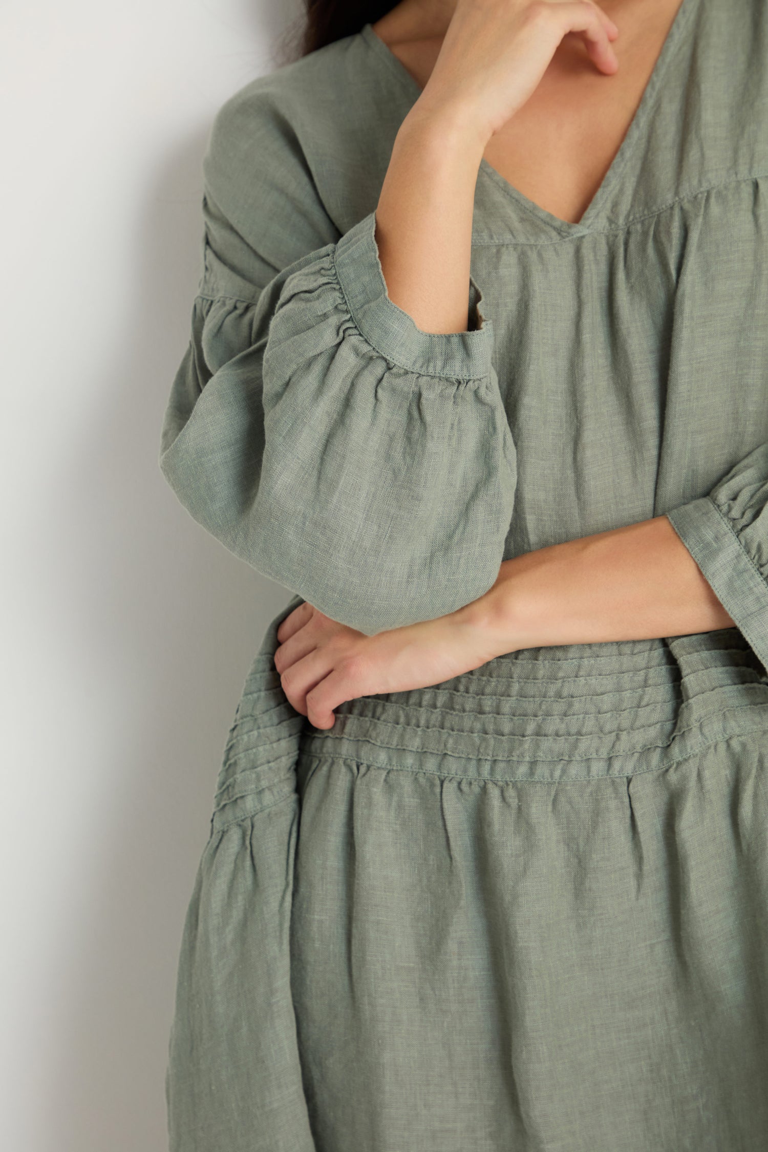 A person is shown from the shoulders down, wearing the Easy Fit V-Neck Sage Linen Dress with puffed sleeves, standing against a plain light background.