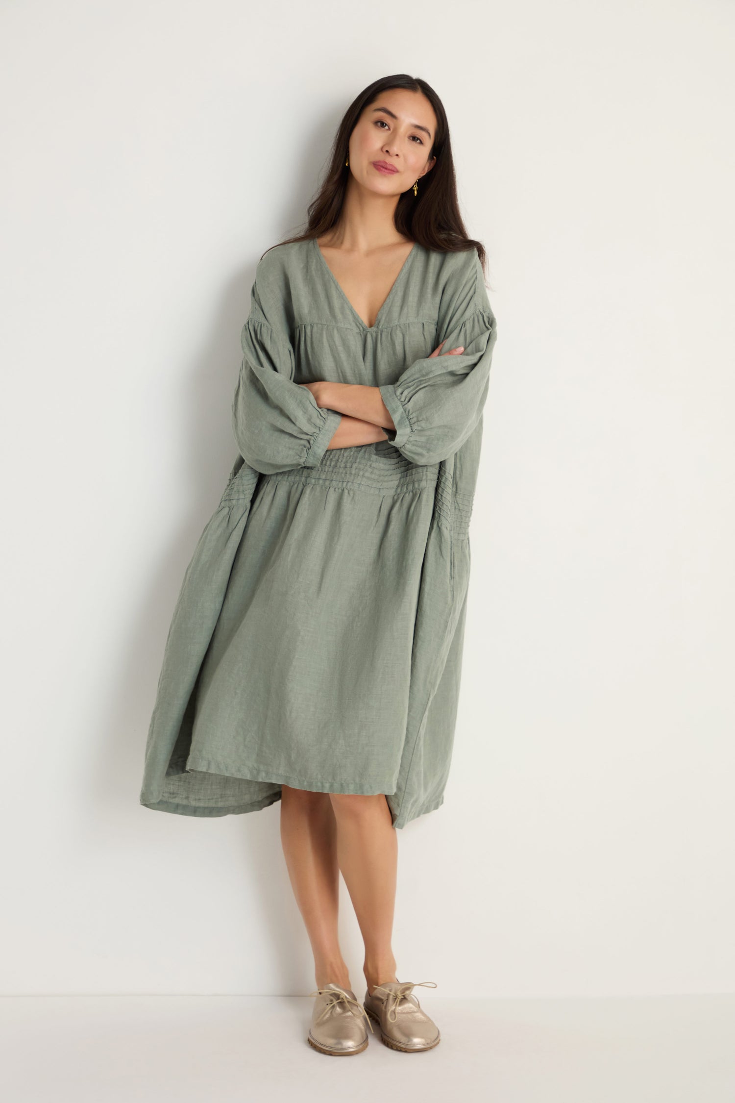 A woman with a neutral expression stands against a white wall, arms crossed, wearing the Easy Fit V-Neck Sage Linen Dress and beige shoes.