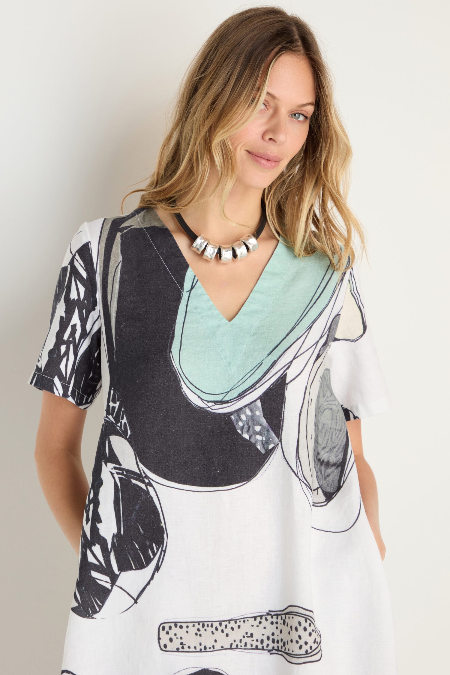 A woman in a Surreal Collage Print Linen Dress with a V-neck and a chunky metallic necklace stands against a plain light-colored background.