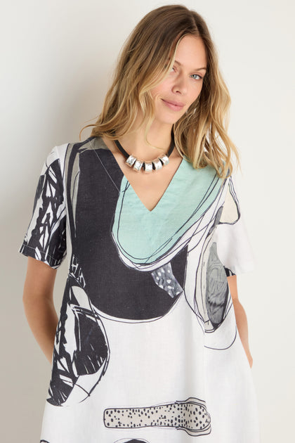A woman in a Surreal Collage Print Linen Dress with a V-neck and a chunky metallic necklace stands against a plain light-colored background.