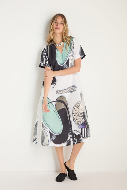 A woman wears the Surreal Collage Print Linen Dress, a loose-fitting, short-sleeve linen dress with abstract black, white, and teal prints, styled with a statement necklace and black shoes against a plain white background.