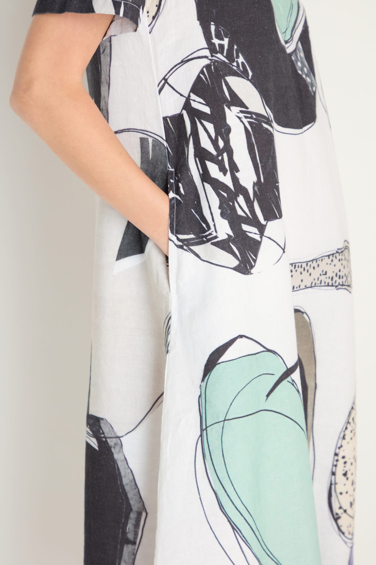 A person wearing the Surreal Collage Print Linen Dress, featuring short sleeves and an abstract black, green, and beige print, stands with one hand in the side pocket.