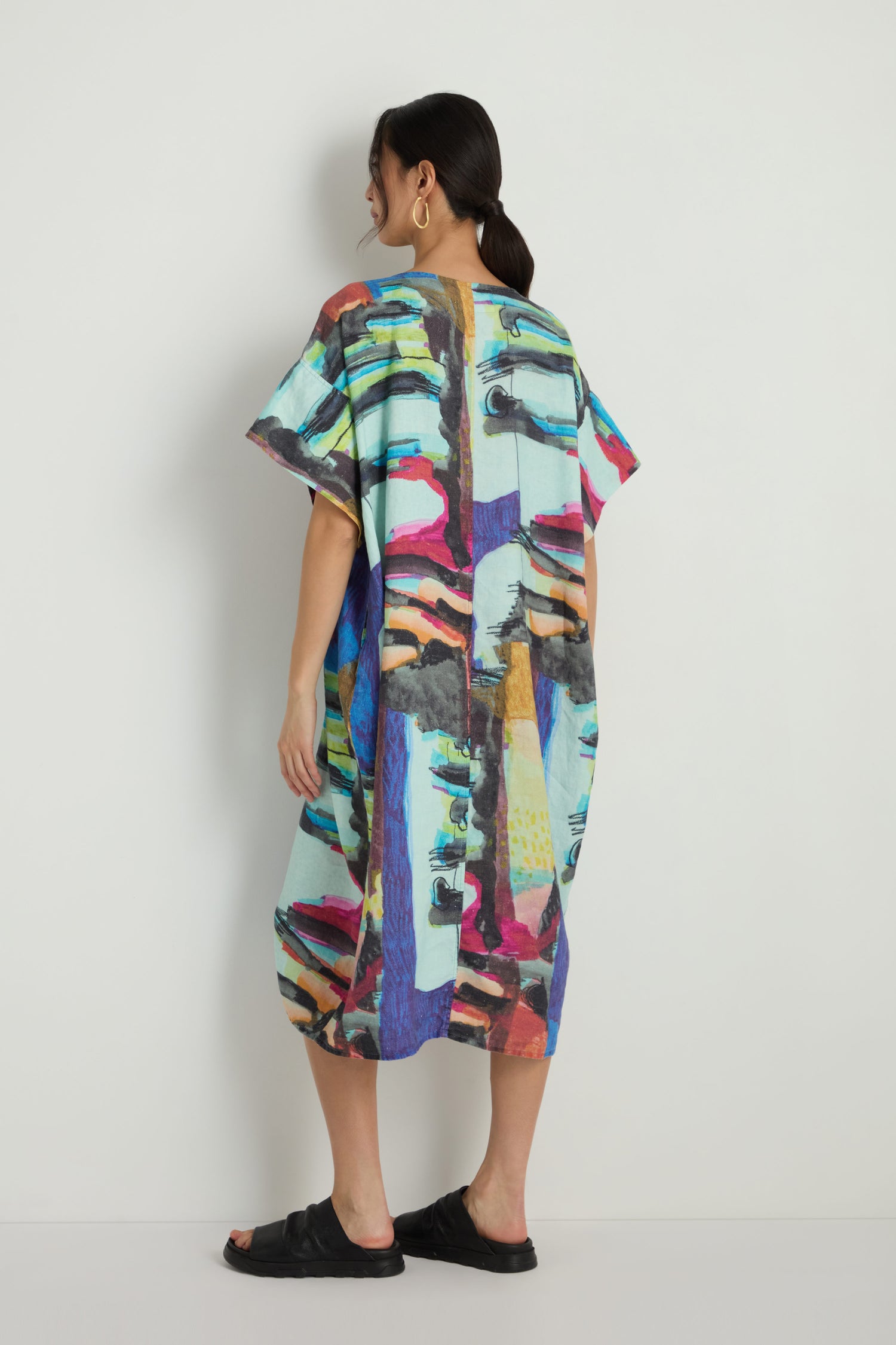 A person stands facing away, wearing the Abstract Print Easy Dress with a colorful abstract pattern and black sandals, set against a plain white background.