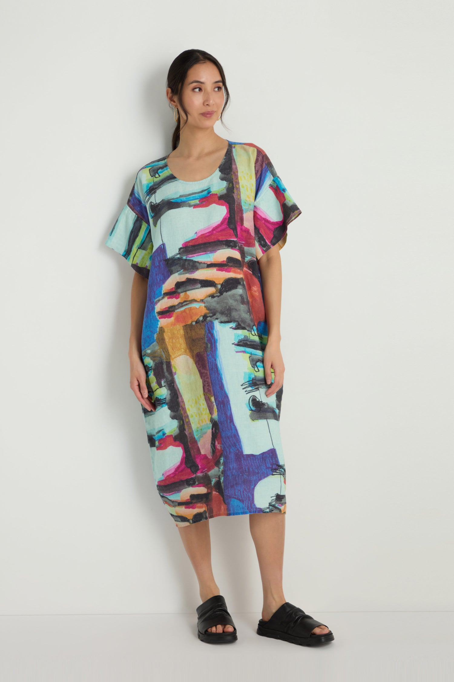 A woman stands against a plain wall wearing the Abstract Print Easy Dress—a loose, knee-length linen dress with a relaxed silhouette and vibrant, eye-catching abstract pattern.