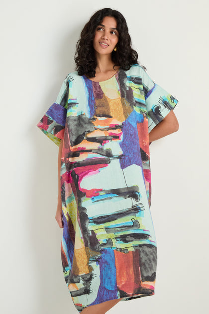 A woman stands against a plain wall wearing the Abstract Print Easy Dress—a loose, knee-length linen dress with a relaxed silhouette and vibrant, eye-catching abstract pattern.