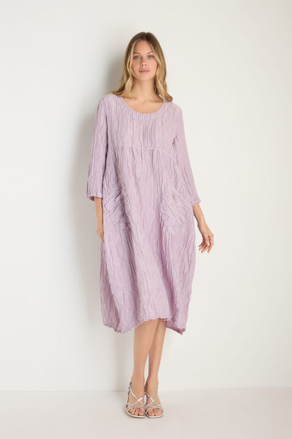 A woman stands against a plain background wearing the Silk Linen Lilac Patch Pocket Dress—a loose, textured dress with three-quarter sleeves and side pockets—paired with strappy sandals.