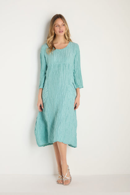 Woman stands against a white wall, wearing a loose-fitting Silk Linen Mint Patch Pocket Dress with three-quarter sleeves in light blue and white strappy sandals.