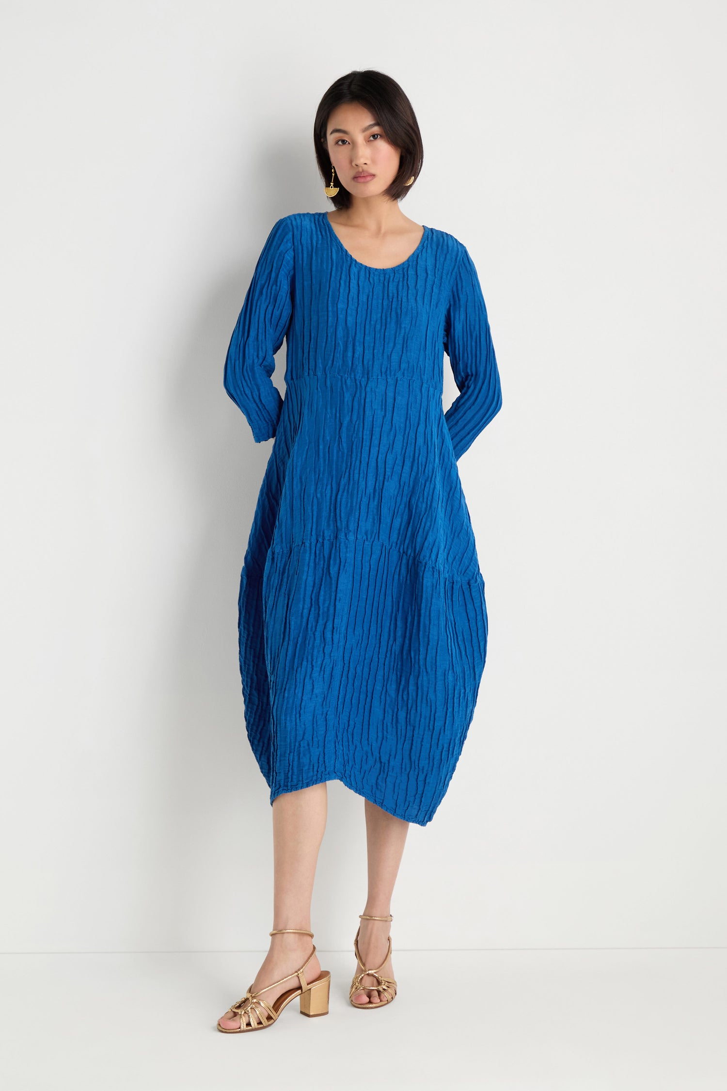 A woman stands against a white wall wearing the Silk Linen Bubble Dress and gold strappy block-heeled sandals.