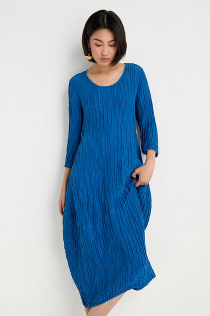 A woman models the Silk Linen Bubble Dress in cobalt, featuring a textured weave, as she stands against a plain white background with one hand in her pocket and looks down.