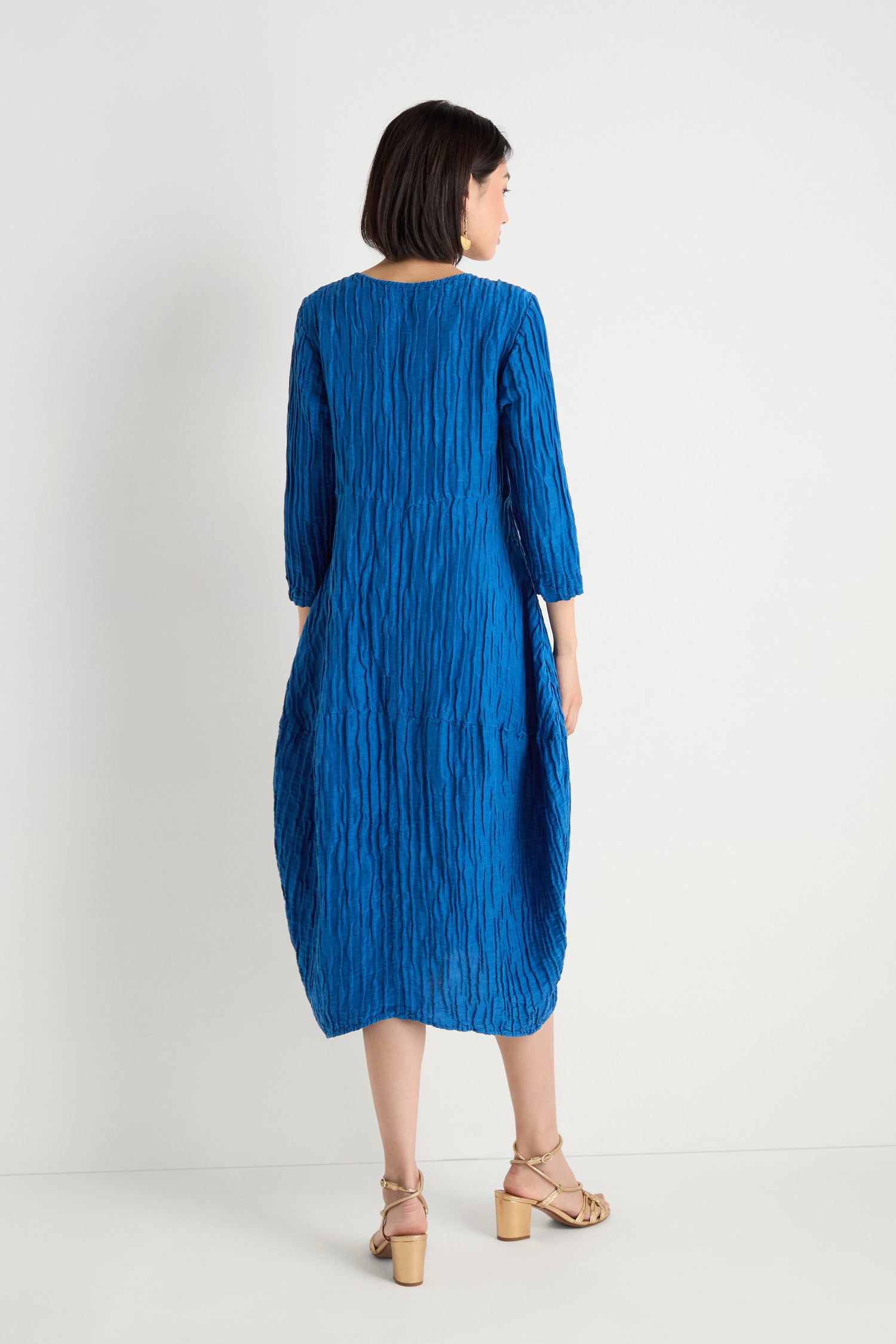 A woman, back turned, wears the Silk Linen Bubble Dress in cobalt with a textured weave and beige heeled sandals against a plain white background.