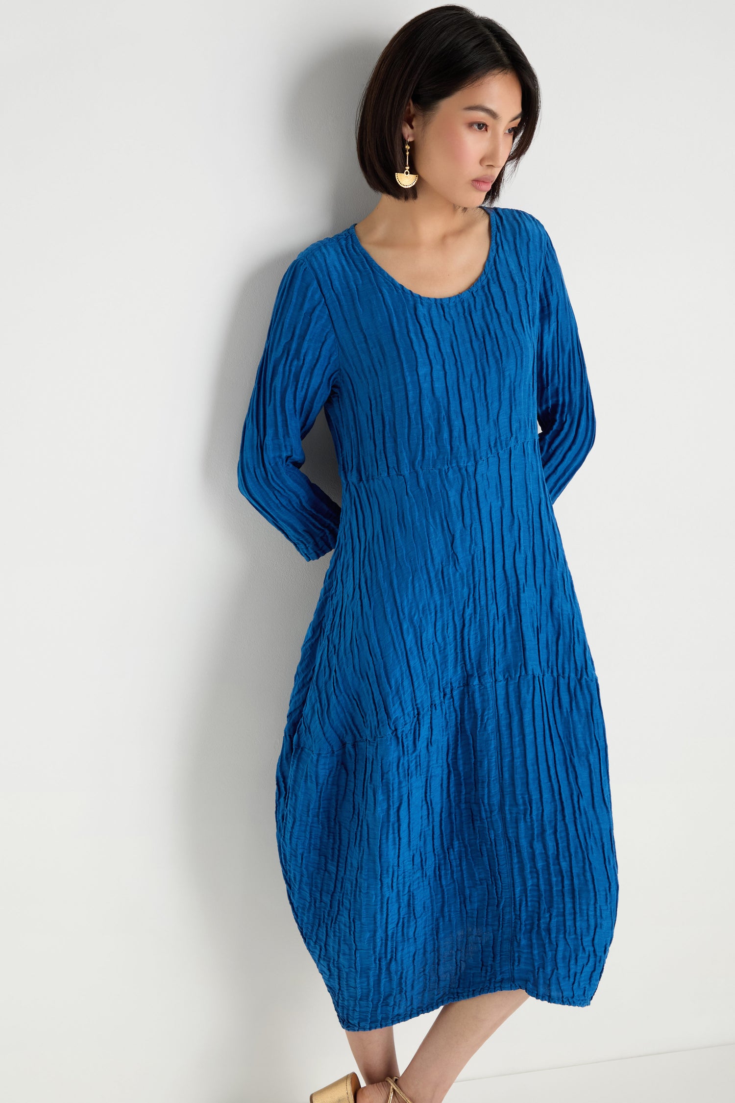 A woman stands against a white wall wearing the Silk Linen Bubble Dress in cobalt with a textured weave and gold earrings, her hands behind her back.