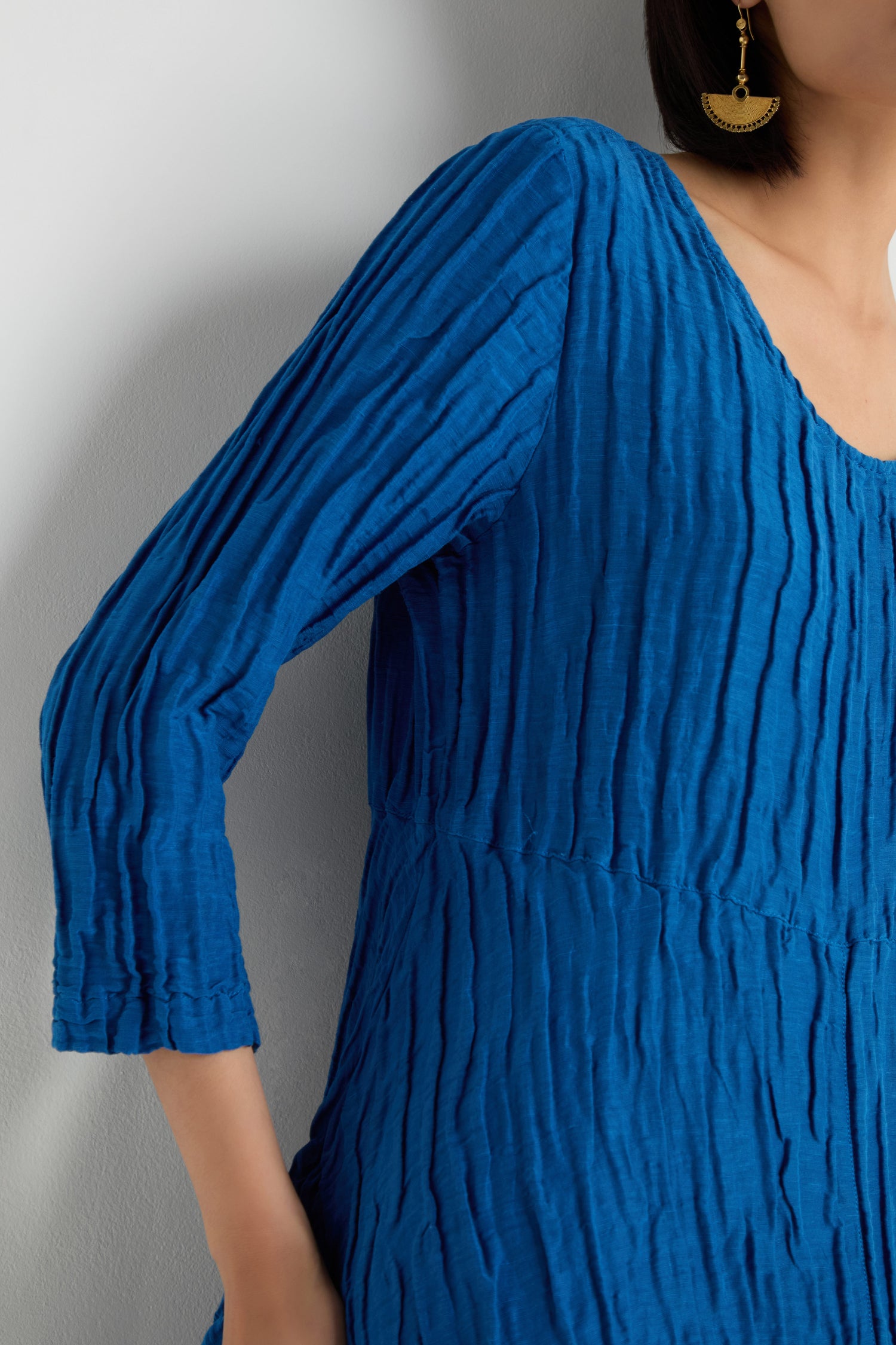 A person wearing the Silk Linen Bubble Dress in cobalt blue with a crinkled, textured weave and gold earrings stands against a light-colored wall.