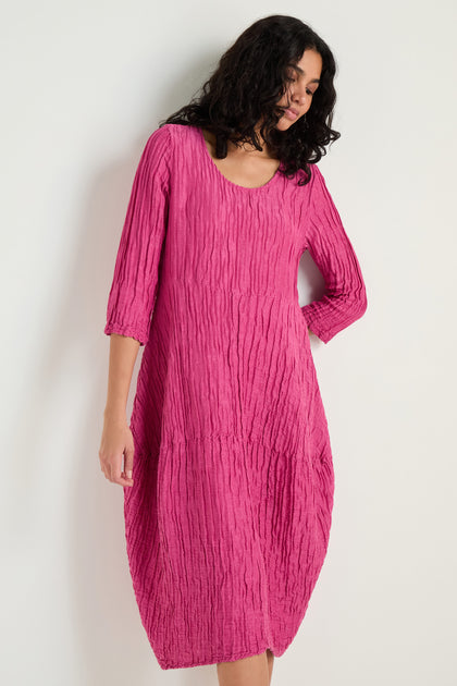 A woman wears the Silk Linen Bubble Dress—a textured weave, knee-length fuchsia dress with three-quarter sleeves—standing against a plain white background.