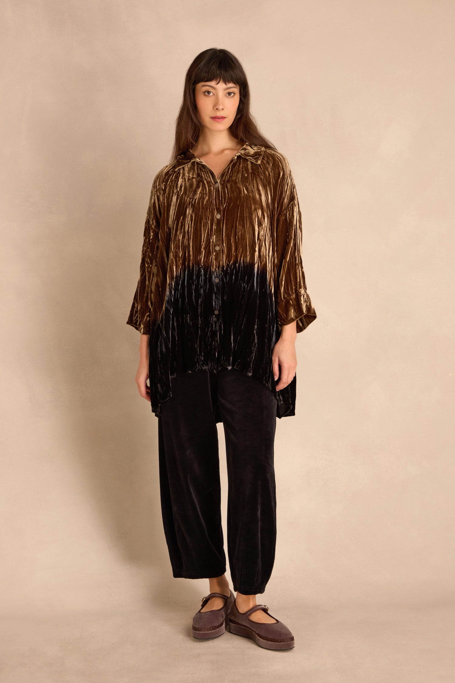 A woman stands against a beige background wearing the Olive Silk Velvet Ombre Shirt with black velvet pants and brown platform shoes.