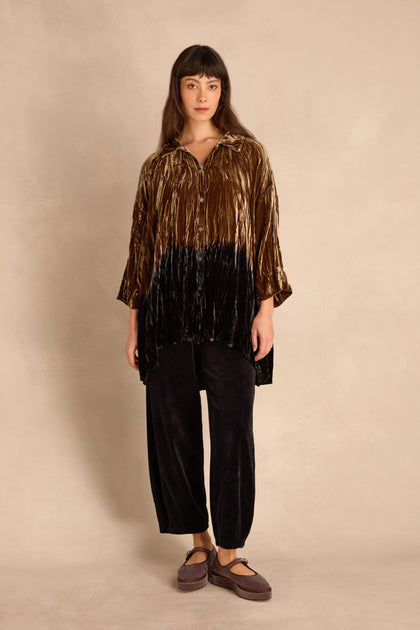 A woman stands against a beige background wearing the Olive Silk Velvet Ombre Shirt with black velvet pants and brown platform shoes.