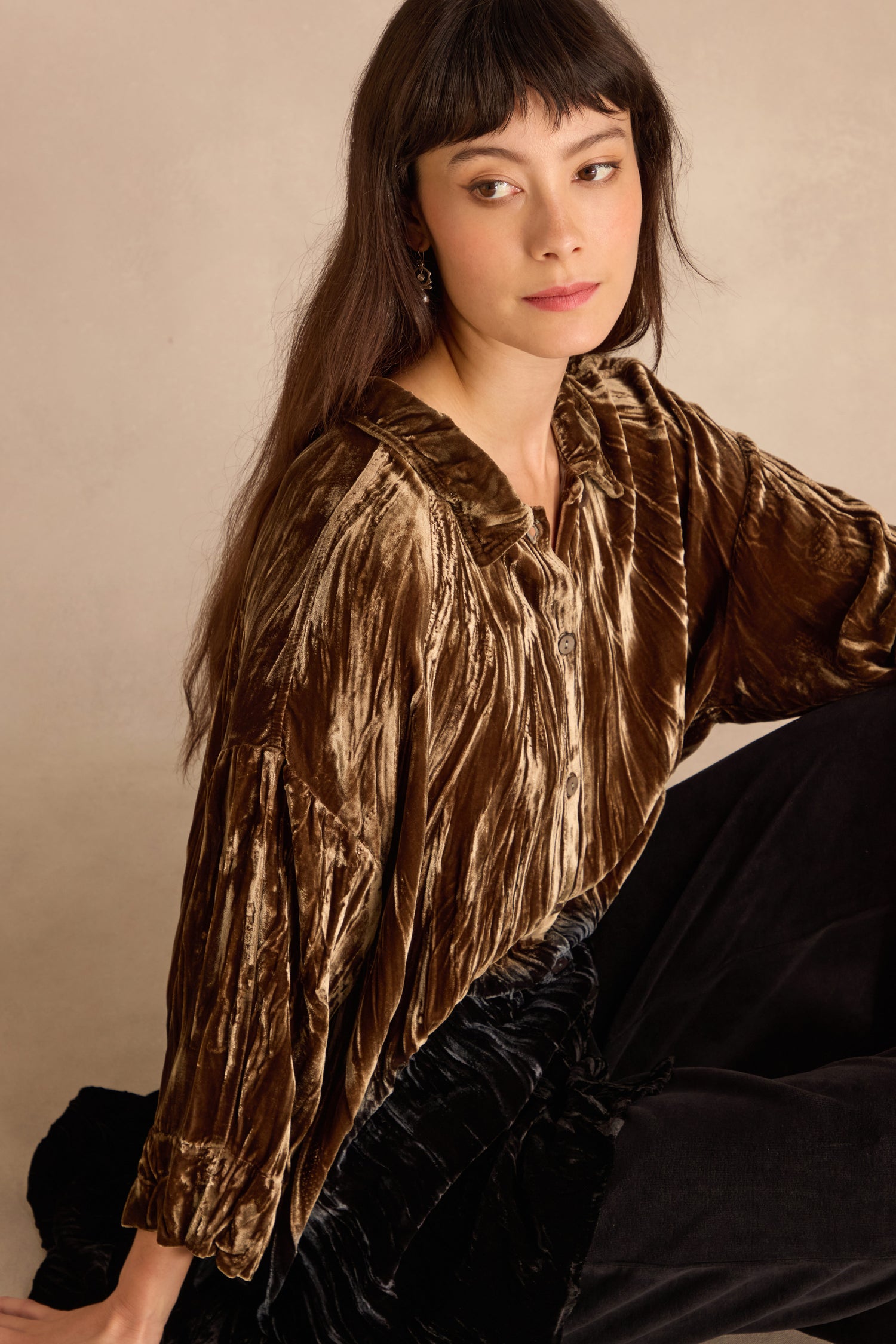 A woman with long dark hair and bangs wears the Olive Silk Velvet Ombre Shirt and black pants, sitting with a neutral expression against a beige background.