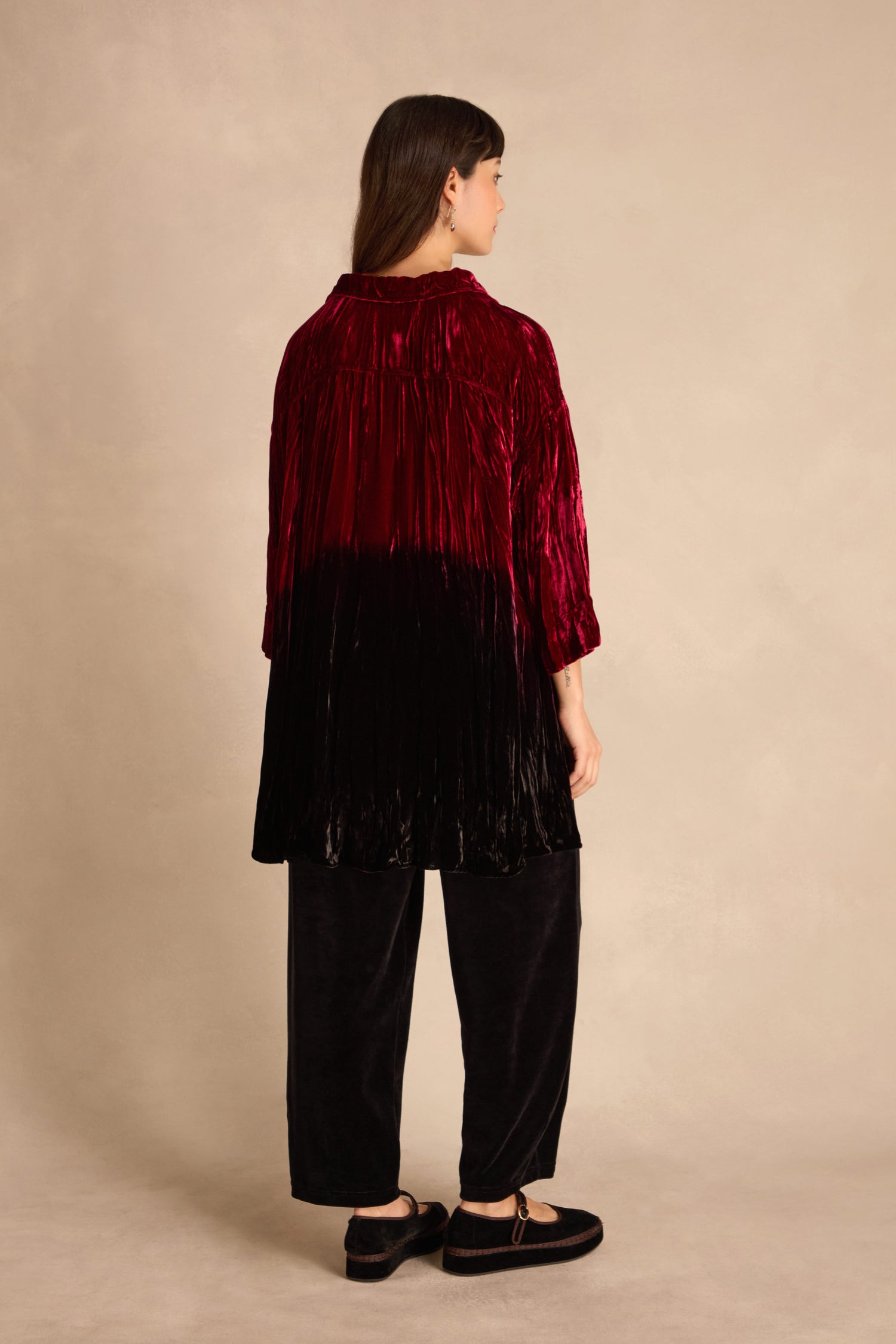 A woman faces away, wearing the Ruby Silk Velvet Ombre Shirt in red-to-black pleats with black wide-leg pants and shoes, set against a neutral background.