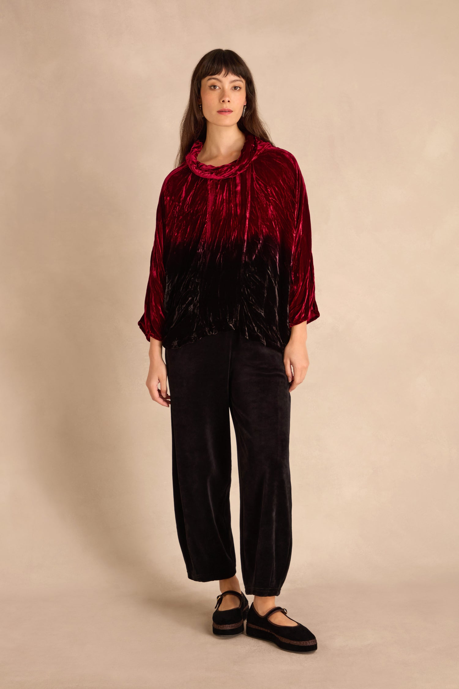 Woman stands against a beige backdrop wearing the Ruby Silk Velvet Ombre Cowl Neck Top, black crushed velvet wide-leg pants, and black shoes.