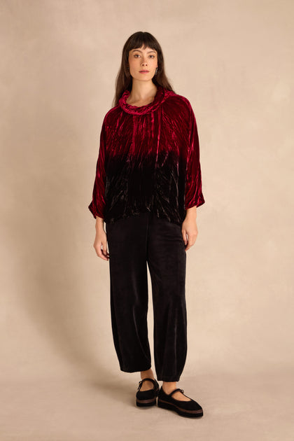 Woman stands against a beige backdrop wearing the Ruby Silk Velvet Ombre Cowl Neck Top, black crushed velvet wide-leg pants, and black shoes.