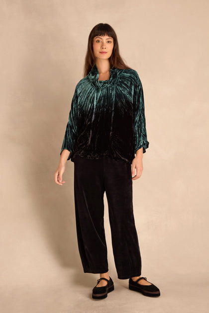 Woman standing against a beige background, wearing the Turquoise Silk Velvet Ombre Cowl Neck Top with black wide-legged pants and black platform shoes.